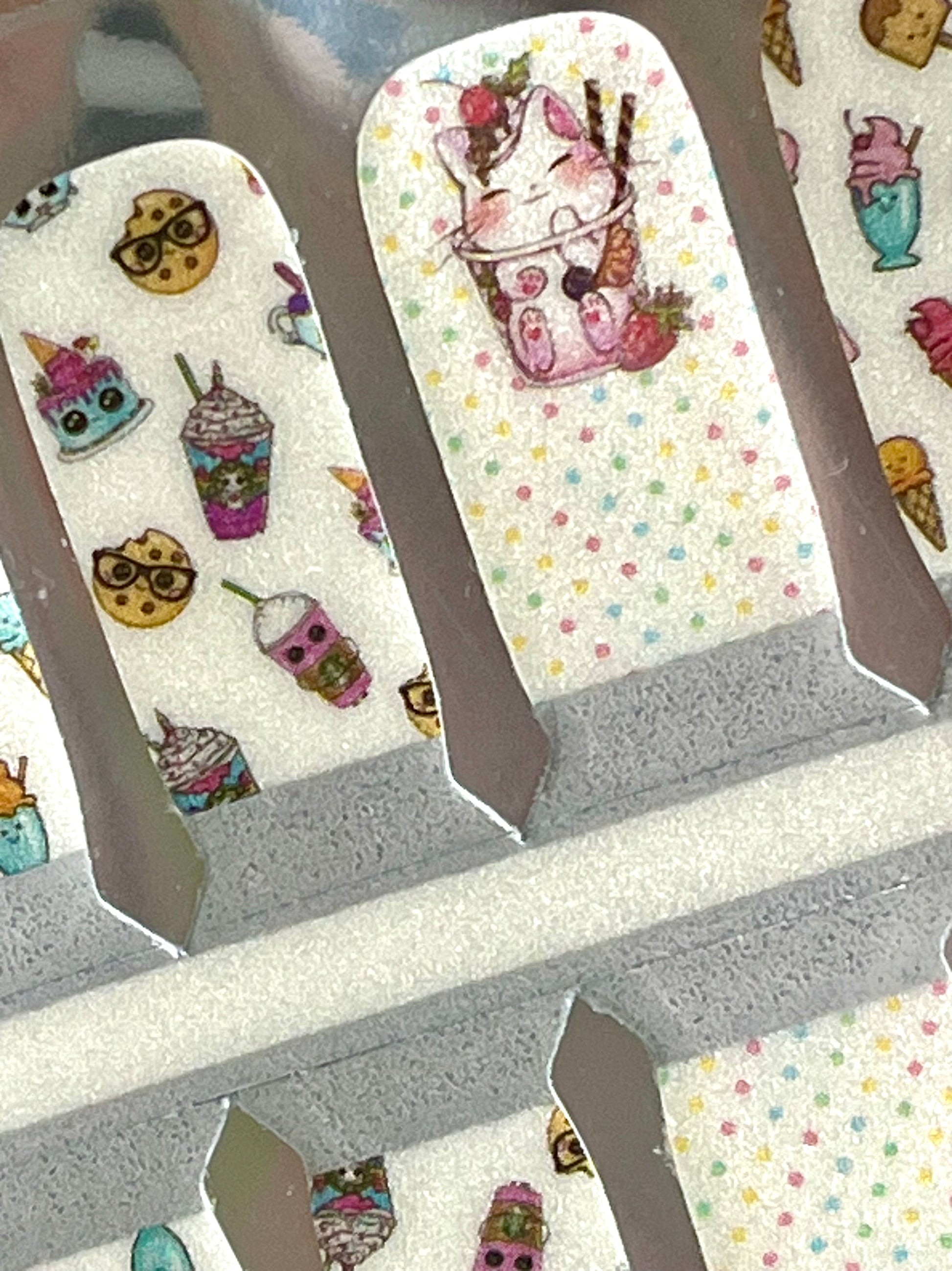 Cute dessert-themed nail wraps featuring cartoon cookies, milkshakes, cakes, and a cat in a boba tea cup on a polka dot background.